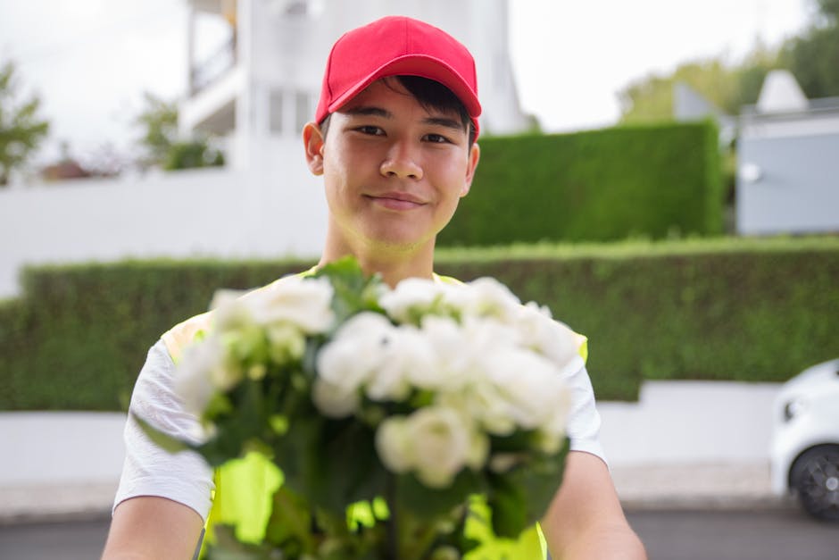 A young delivery person wearing a red cap and high-visibility vest smiling while holding a vibrant bouquet of white flowers with green leaves. The bouquet features fresh, blooming petals and lush stem